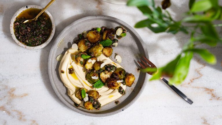 Des choux de Bruxelles grillés sur du houmous, servis dans une assiette. À côté se trouve un petit bol de vinaigrette au sumac. 