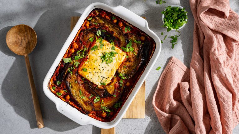 Gebackener Feta mit Aubergine in Tomatensauce in einer weißen Auflaufform auf grauem Untergrund. Es liegt ein rosa Tuch daneben.