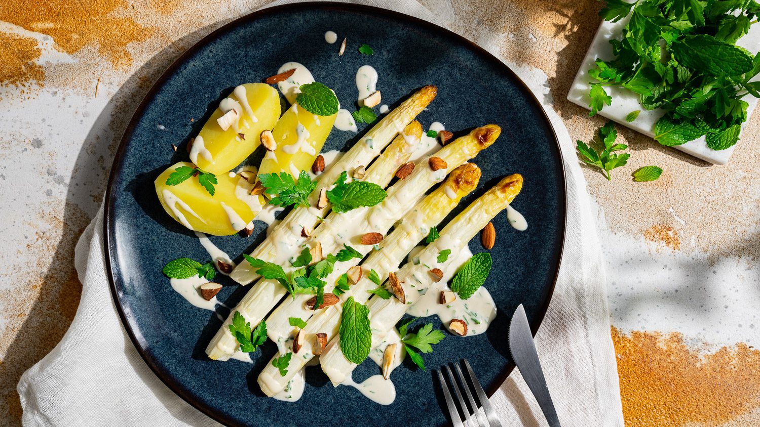 spargel mit tahinisauce, kartoffeln und kräutern auf einem blauen teler angerichtet.