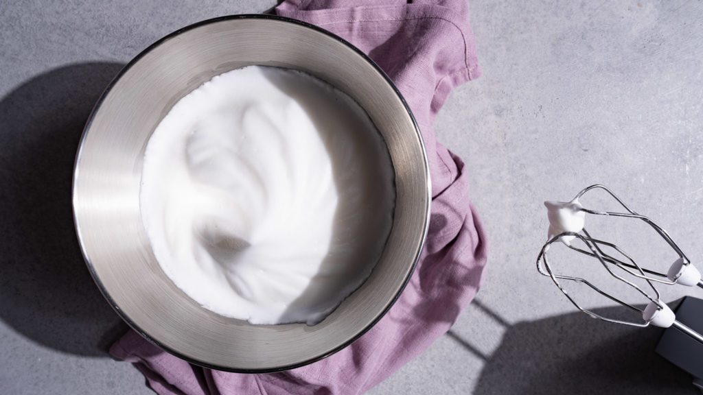 Veganer Eischnee aus Aquafaba in einer Rührschüssel. Daneben steht ein Handmixer.