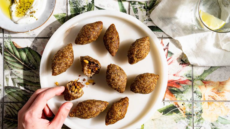 Classic kibbeh croquettes filled with minced meat are served on a round marble plate. A kibbeh is cut in half and gripped by one hand.