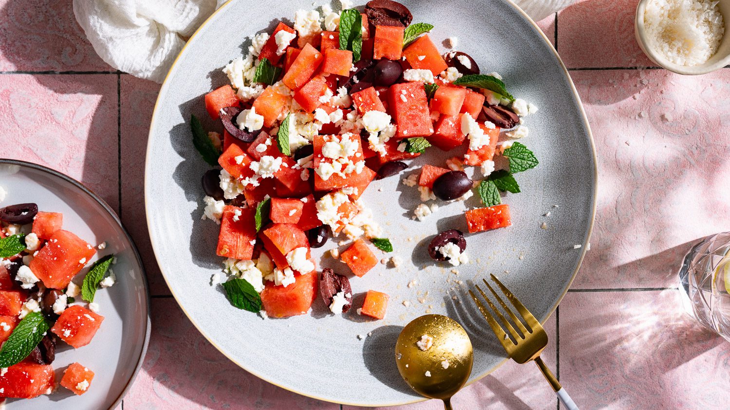 Ensalada de melón con queso feta, menta y aceitunas servida en un plato grande. El fondo es de color rosa.