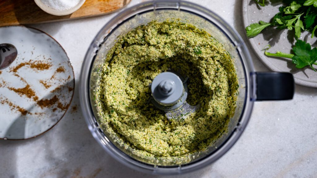 Auf einem hellen Marmoruntergrund steht ein geöffneter Mixer mit den zerkleinerten Zutaten für Falafel darin.