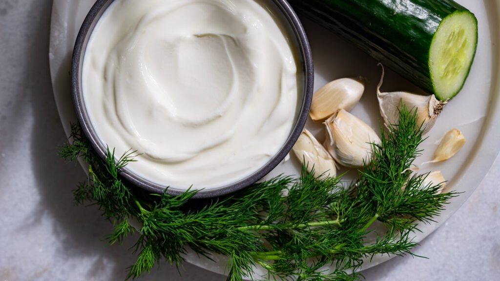 Greek cream yogurt, dill, cucumber and garlic cloves on a platter.