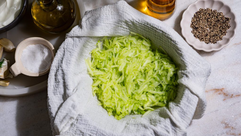 A grated cucumber is in a straining cloth. Around them are other ingredients for tzatziki.