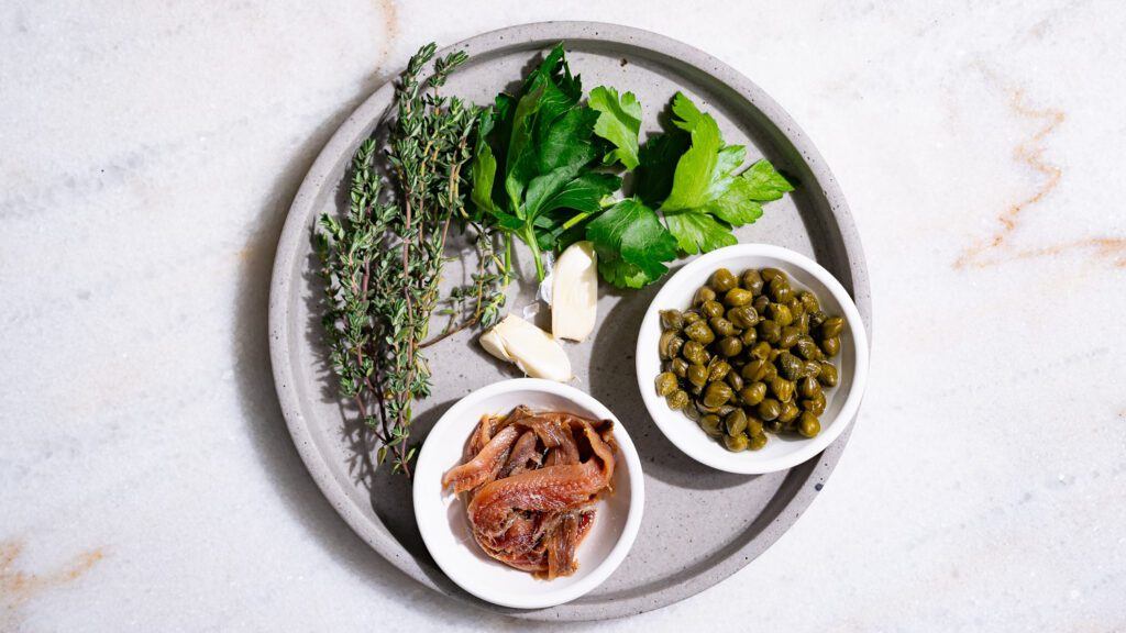 Des herbes, des câpres, de l'ail et des filets d'anchois sur une assiette grise.  