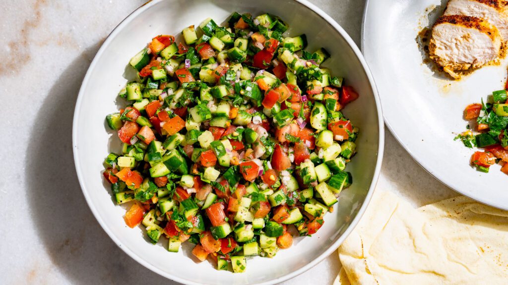 Ensalada Shirazi servida en un cuenco. A su lado hay un plato de ensalada y pollo.