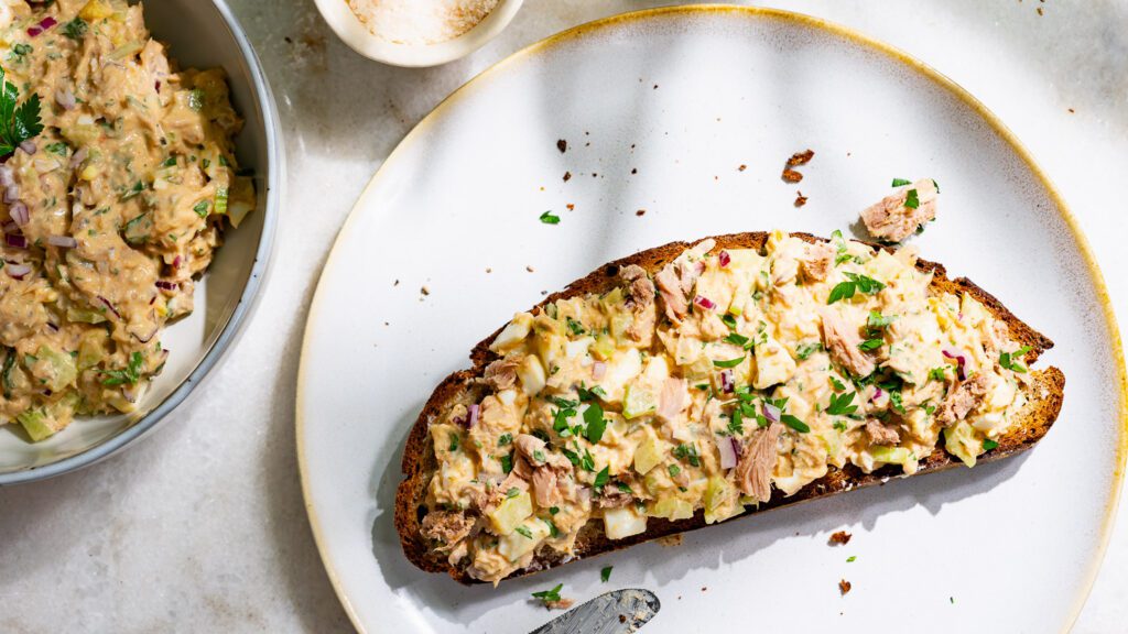 Thunfischsalat auf einer Scheibe Brot verstrichen. Daneben steht eine Schüssel mit mehr Thunfischsalat