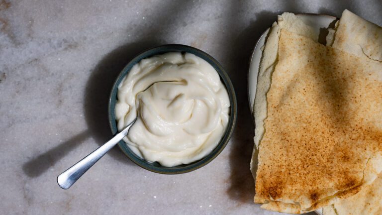 Toum mit einem Löffel in einer Schüssel serviert. Daneben liegt libanesisches Fladenbrot.