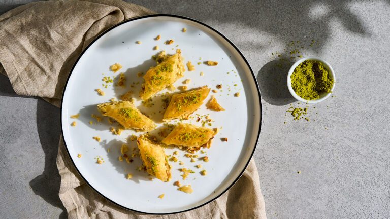 Origen del baklava: Baklava servido en un plato. A su lado hay un pequeño cuenco con pistachos. 