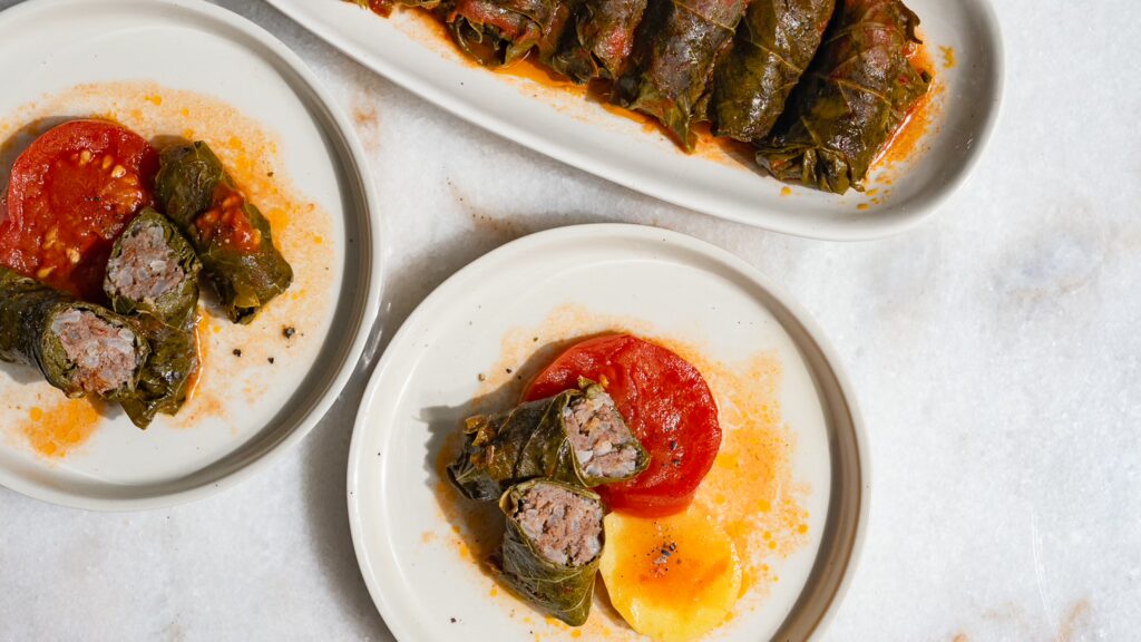 Hojas de parra rellenas de carne picada servidas en dos platos pequeños con tomate. Al fondo, una fuente con hojas de parra rellenas.